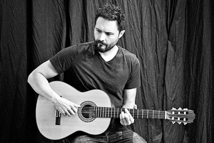 Andrew Sisk playing a classical guitar in front of a dark curtain